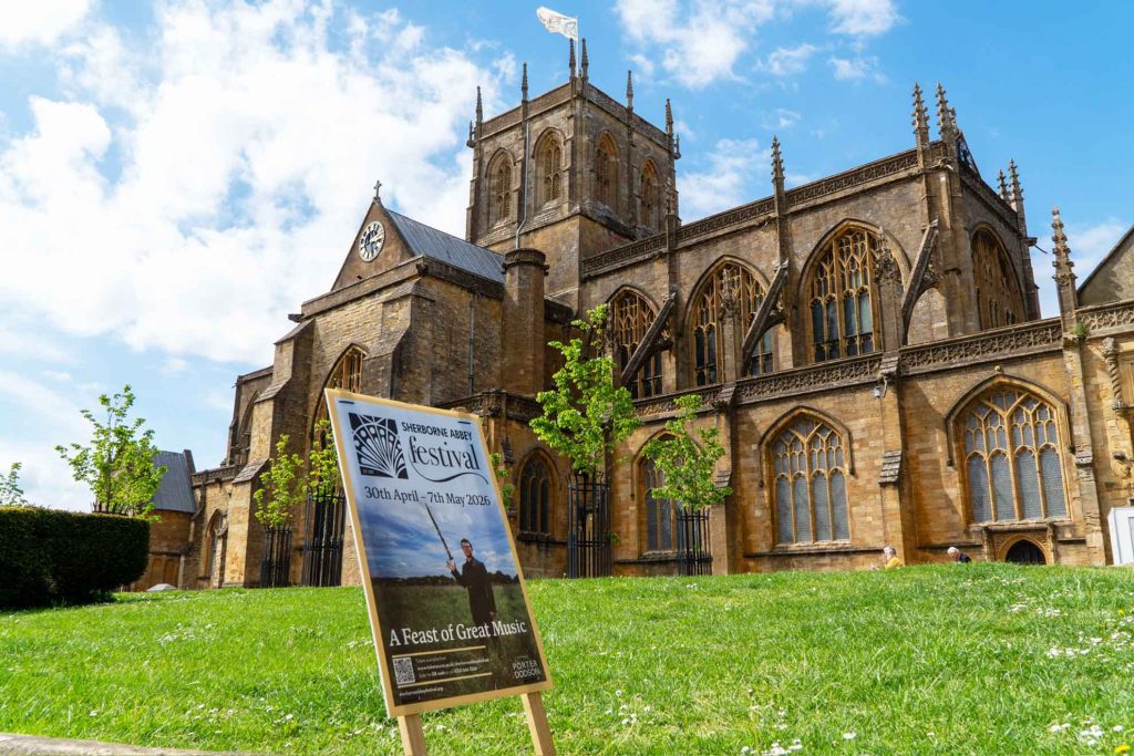 Sherborne Abbey on a sunny day, with Festival poster in front.